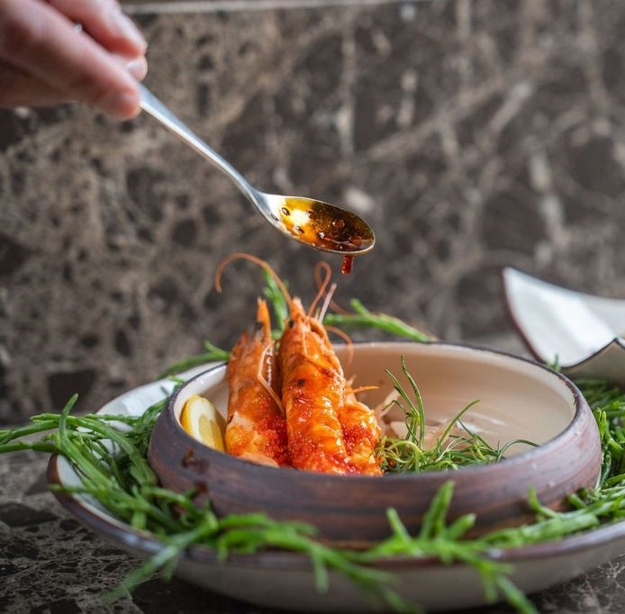 Hand holding a spoon drizzling sauce over a dish of shrimp.